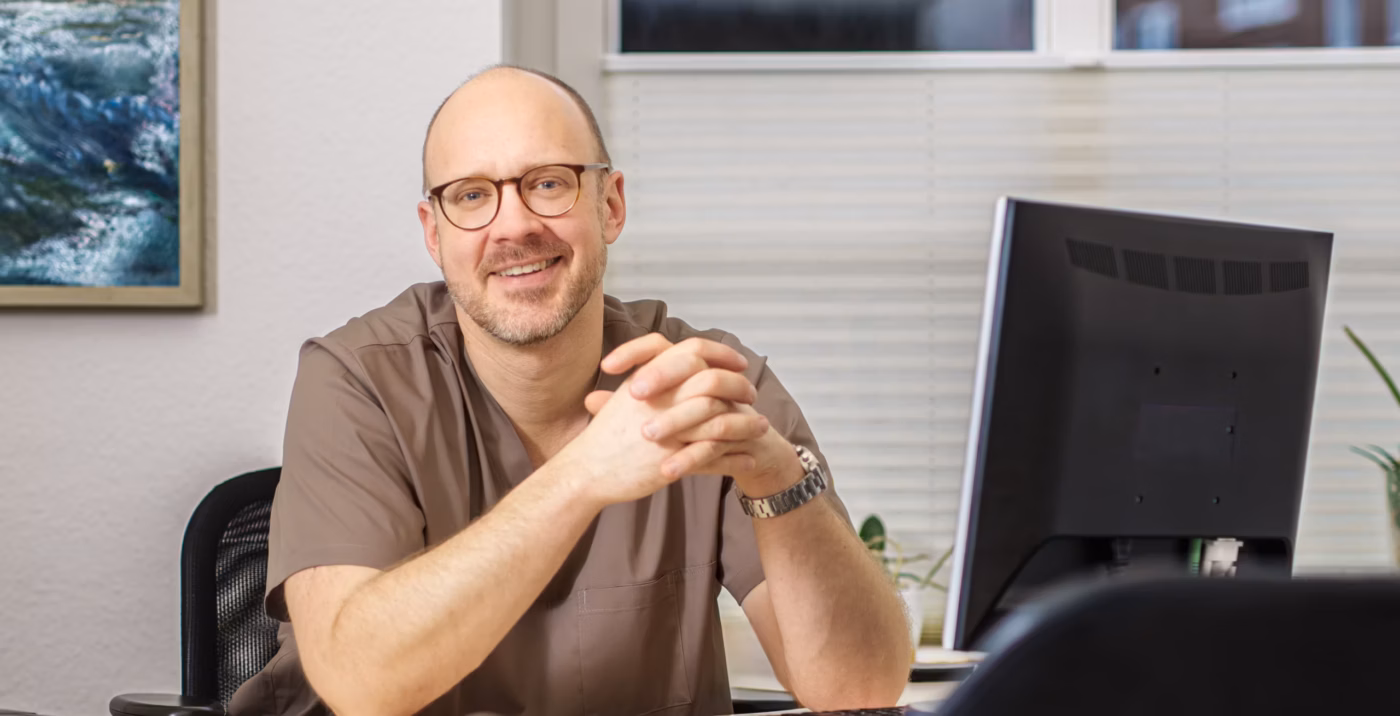 Dr. med. Reitz sitzt lächelnd an seinem Schreibtisch in der Praxis